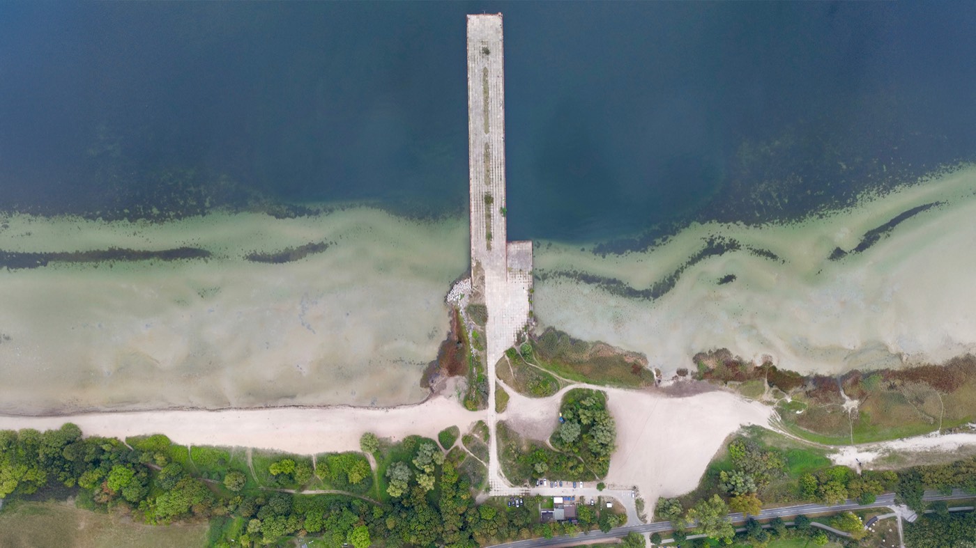 Senkrechte Luftaufnahme: der Anleger ragt schnurgerade in das türkisgrüne Wasser der Wiek, davor Sandstrand, Bäume und ein Parkplatz.